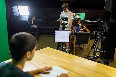 Over the shoulder image of student at a table. He is facing a student operating a camera with a teleprompter in front of it. 